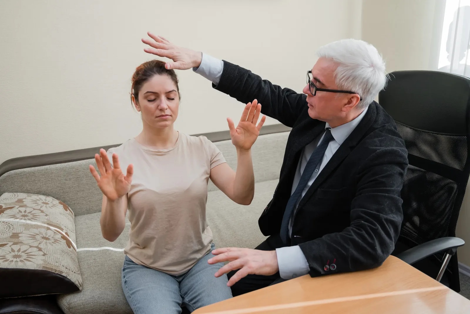 A hypnotherapy session in progress: a practitioner sits beside a client who is seated with her eyes closed and hands raised.